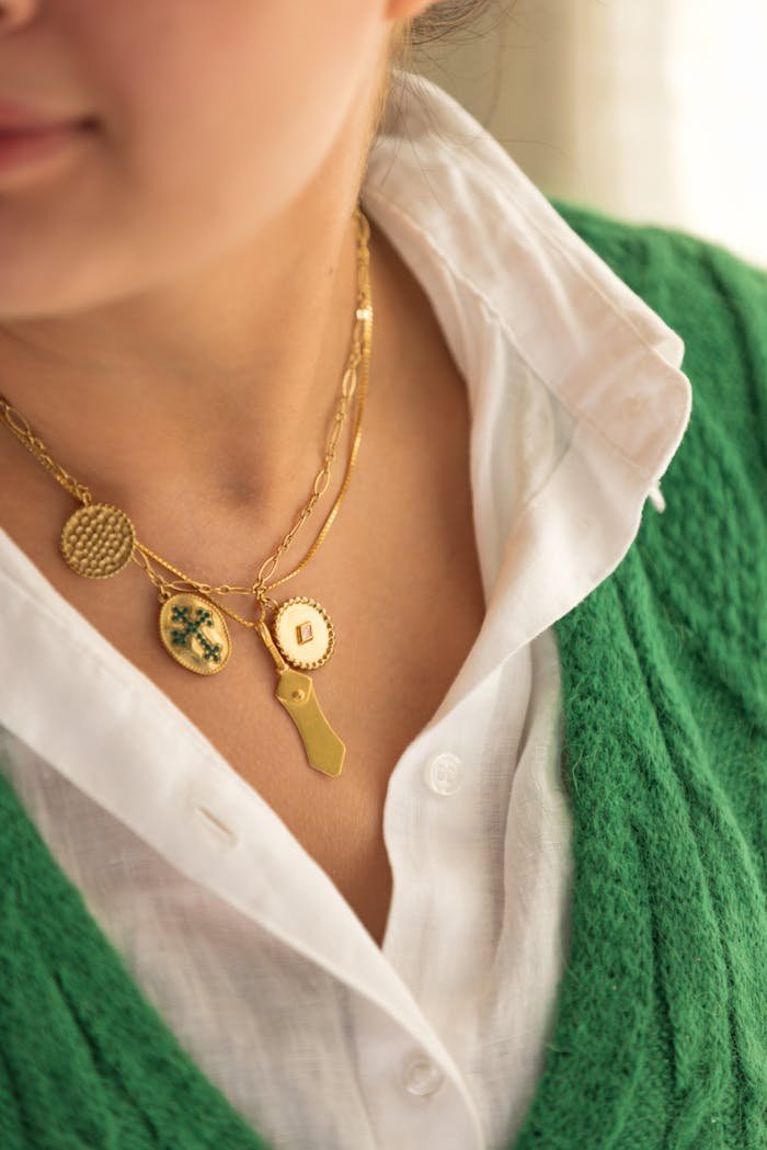Close-up of a woman's gold necklace with charms over a white shirt and green sweater, stylish and elegant.